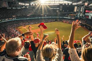 A lively crowd at a baseball game cheers enthusiastically, waving red towels in a sunlit stadium filled with fans. Energy and excitement abound.