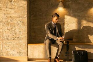 A man in a suit works intently on a laptop, seated in a warmly lit, modern space, with a briefcase nearby.