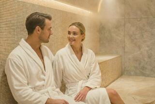 A man and woman in white robes sit comfortably in a serene steam room, sharing smiles and enjoying their relaxing moment together.