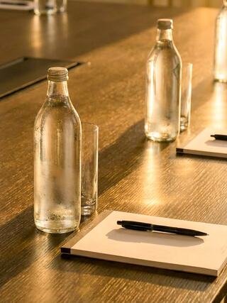 Clear glass bottles and glasses of water sit beside notepads and a pen on a polished wooden table, illuminated by warm light.