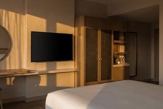 A stylish hotel room features warm sunlight, a flat-screen TV, a minimalist desk, and a wooden cabinetry with refreshments.