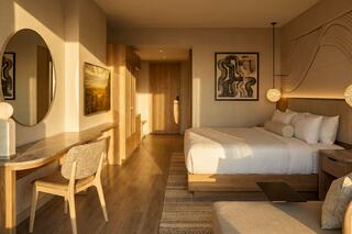 A warm, modern hotel room features a cozy bed, wooden accents, a round mirror, and soft lighting, creating an inviting atmosphere.