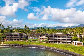 Tropical resort featuring modern buildings amid lush palm trees, a pool area, and scenic mountains under a bright blue sky.