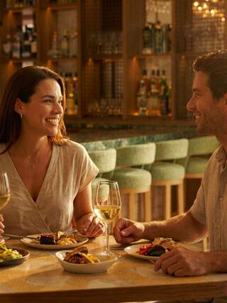 A couple enjoys a cozy dinner in a warmly lit setting, sharing smiles and delicious food while sipping wine at a stylish bar.