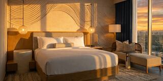 A beautifully designed hotel room features a large bed, stylish furnishings, and a stunning sunset view through floor-to-ceiling windows.