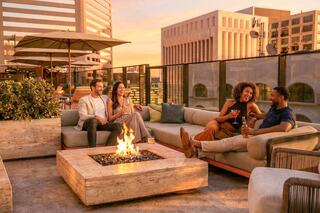 A group of friends enjoys drinks around a cozy rooftop fire pit, with stunning city views at sunset. Laughter and warmth fill the scene.