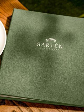 A green menu with the name "SARTÉN KITCHEN & BAR" embossed, resting on a wooden table near a plate. Natural light creates a soft shadow.