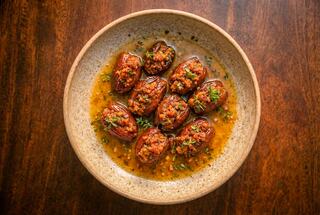 A bowl of stuffed dates, drizzled with a flavorful sauce and garnished with herbs, sits on a wooden surface, showcasing a delicious dish.