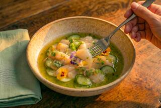 A bowl of fresh seafood, garnished with herbs and edible flowers, with a fork poised to take a bite, set on a wooden table.
