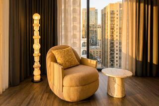A cozy, modern chair with a decorative pillow sits next to a marble side table and a unique lamp, framed by cityscape views.