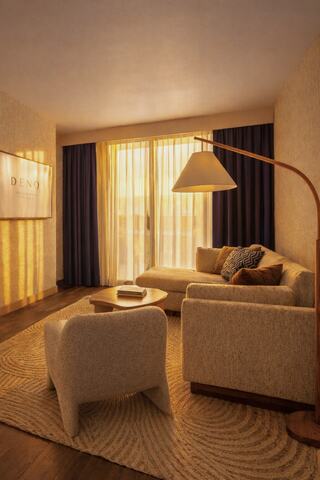 A cozy living space features a soft sofa, stylish chairs, and warm lighting, creating an inviting atmosphere with large windows and curtains.