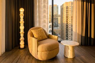 A cozy armchair with a decorative pillow sits beside a marble table and a unique floor lamp, illuminated by warm, natural light from large windows.