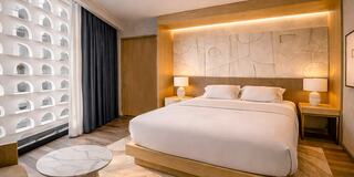 A modern, warmly lit bedroom features a large bed, stylish decor, and large windows with a view, creating a cozy and inviting atmosphere.