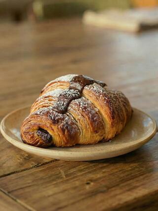 A flaky, golden-brown pastry with chocolate drizzle sits on a rustic plate, dusted with powdered sugar, against a wooden table.