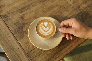 A cup of latte art featuring a leaf design rests on a saucer, with a hand poised to enjoy it on a rustic wooden table.