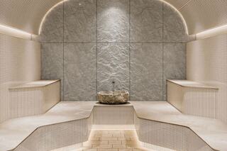 A serene spa space features smooth stone walls, minimalist seating, and a central stone fountain, illuminated by soft ambient lighting.