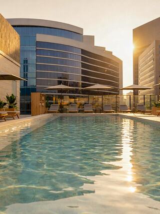 A contemporary rooftop pool area featuring lounge chairs and umbrellas, surrounded by sleek, modern buildings under a warm, golden light.