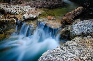 A gentle stream flows over smooth rocks, creating a tranquil scene with soft water currents and natural greenery in the background.