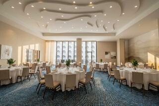 A bright, elegant dining room features round tables set for a formal event, with modern decor and large windows allowing natural light.