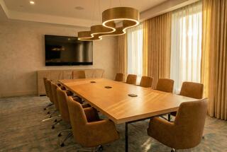 A modern conference room featuring a large wooden table, comfortable brown chairs, and warm lighting, with a TV and large windows.