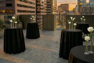 Rooftop setting featuring elegantly set tables with black cloths and floral centerpieces, with a city skyline at sunset in the background.