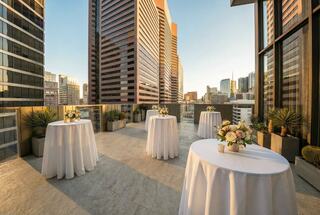 A stylish rooftop terrace features elegant tables adorned with flowers, overlooking a skyline of modern skyscrapers at sunset.