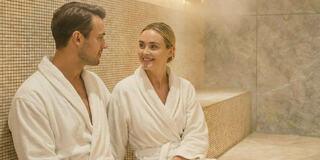 A man and woman in white robes sit in a steam room, enjoying each other's company and sharing smiles in a relaxed atmosphere.
