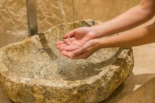 Water flows gently from a modern faucet into a textured stone basin, as hands cup the water beneath, creating a serene and natural atmosphere.