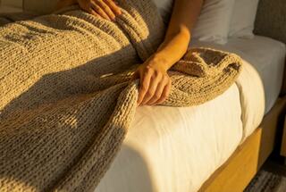 A cozy bed scene with warm sunlight highlighting a hand gently holding a textured blanket, evoking a sense of comfort and relaxation.