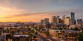 A vibrant city skyline at sunset, featuring tall buildings and palm trees, with mountains in the background under a colorful sky.