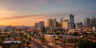 A vibrant cityscape at sunset, featuring modern skyscrapers, palm trees, and distant mountains, creating a warm, atmospheric glow.