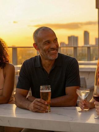 A smiling man enjoys drinks with friends at a rooftop bar during sunset, surrounded by a vibrant city skyline.