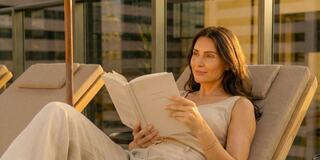 A woman relaxes on a balcony, reading a book in the sunlight, dressed in light clothing, with a serene expression. Modern city skyline in the background.