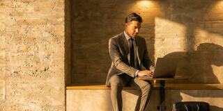 A man in a suit works intently on a laptop, seated against a textured wall, illuminated by warm, natural light.