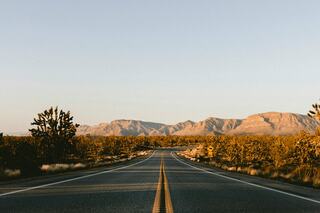 A long, empty road stretches toward distant mountains under a clear sky, framed by sparse vegetation and desert landscapes.