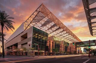 A striking modern building with a large, angular roof, surrounded by palm trees, set against a vibrant sunset sky.