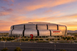 A modern stadium with a sleek, metallic exterior, set against a colorful sunset, surrounded by a parking lot and desert landscape.