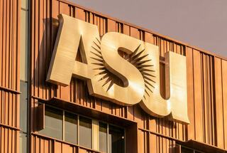 A bold, metallic sign displays "ASU," enhanced by rays of sunlight, set against a modern, warm-toned building facade.