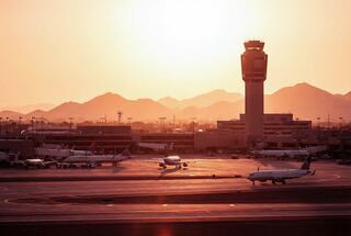An airport at sunset, with planes parked on the tarmac and a control tower silhouetted against a backdrop of mountains.