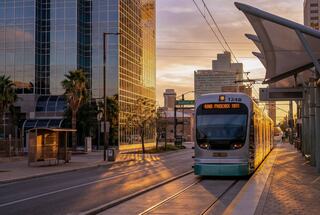 A light rail tram approaches a station at sunset, surrounded by modern buildings and palm trees, casting warm golden hues across the scene.
