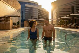 A couple stands in a pool at sunset, surrounded by modern buildings and lounge chairs, enjoying a moment together.