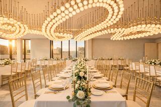 A sophisticated dining space with elegant tables, golden chandeliers, and floral centerpieces, illuminated by warm sunset light.