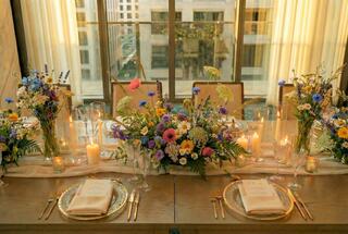 A beautifully arranged dining table adorned with vibrant flowers, candles, and elegant tableware, set against a backdrop of large windows.