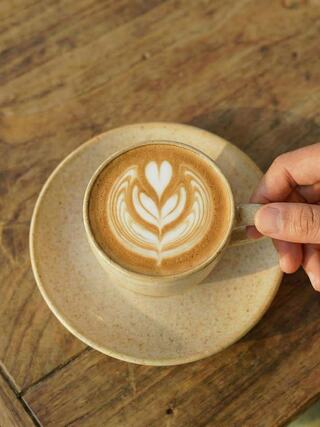 A hand gently holds a latte in a textured cup, showcasing intricate heart-and-leaf latte art, resting on a simple saucer atop a wooden table.