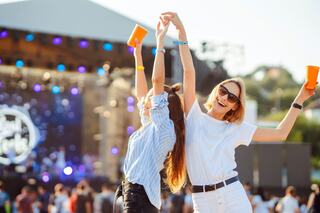Two happy women celebrate at a festival, raising cups and dancing under colorful lights while a crowd enjoys the lively atmosphere.