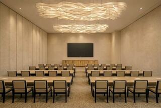 A modern conference room featuring rows of tables and chairs, with a large screen and an elegant, wavy light fixture overhead.