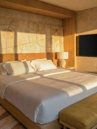 A modern bedroom featuring a large bed with white bedding, warm wooden accents, a stylish lamp, and natural sunlight casting soft shadows.