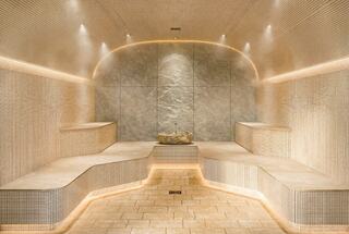 A serene spa space features smooth stone benches, soft lighting, and a central decorative element, enveloped in gentle steam.