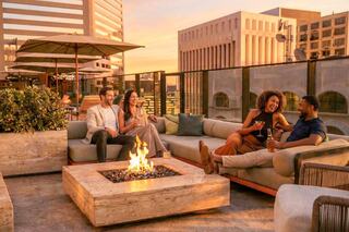 A group of friends enjoys drinks and laughs by a rooftop fire pit, with a stunning sunset backdrop and cityscape beyond.