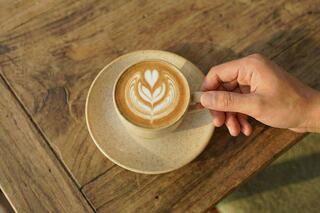 A hand delicately holds a cup of coffee with beautiful latte art, resting on a rustic wooden table. Soft lighting enhances the cozy vibe.